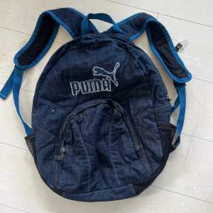 En blå ryggsäck från Puma med justerbara axelremmar och flera fack. Väskan har en dragkedja framtill och är tillverkad i ett jeansliknande material.