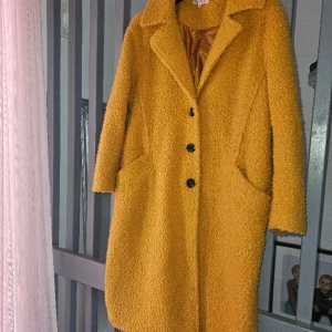 Senapsgul Teddy Kappa - Längre kappa i klassisk rak modell, härligt yttre tyg i Teddy och glansig silke inuti. Fint skick. Passar M-XL beroende på passform man vill ha. Se mått för korrekt bedömning: B 64. L 118. Nypris 799kr