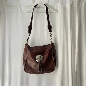 Brun axelremsväska - Brun skinn axelremsväska. Bredd: 34 cm. Djup: 13 cm. Höjd: 32 cm. Den har två fickor innuti med dragkedka, den ena är trasig men funkar ändå som en ficka 💕 