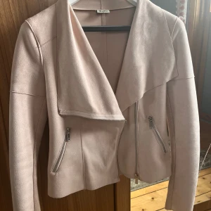 Stilig vårjacka i rosa - Jättefin rosa vårjacka med trendigt snedvridet blixtlås, aldrig tidigare använd!  Elastiskt tyg vid armarna som underlättar rörelse. Den har även en fin krage som passar som såväl till när jackan är öppen som stängd! Silvriga blixtlås som detalj!