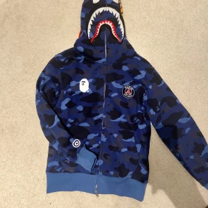 Bape PSG Hoodie  - Bape PSG Zip Hoodie, inte använda, prislappen kvar, bape påsen också kvar. Skriv vid andra frågor. Inte plastig material, det står M men jag skulle säga att det är Stor S  