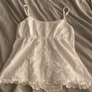 Vit spetslinne - En vit spetslinne med smala axelband och elastisk midja. Linnet har ett broderat mönster och volangdetaljer vid nederkanten.