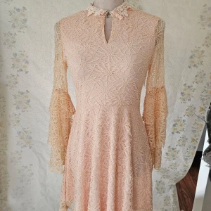 Ljusrosa spetsklänning från Voyelles - En elegant ljusrosa spetsklänning från Voyelles. Klänningen har långa ärmar med volangdetaljer och en hög hals med en liten öppning framtill. Den är tillverkad i ett vackert spetsmaterial och har en dragkedja på sidan. Fläck finns vid dragkedjan som jag ej försökt ta bort.