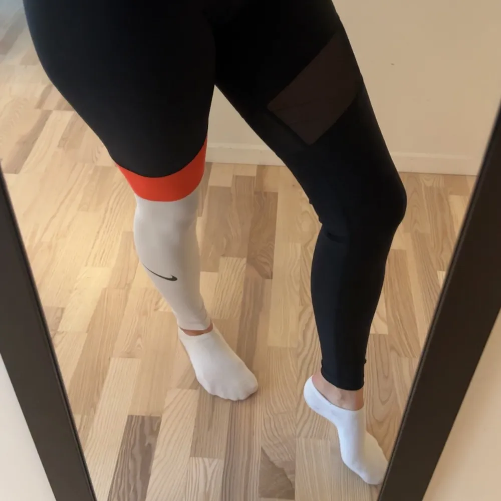 Ett par Nike Dri-Fit leggings i storlek S. Byxorna är huvudsakligen svarta med en vit sektion på vänster ben och en orange detalj vid midjan. De är designade för träning och är tillverkade av ett syntetmaterial som andas.. Jeans & Byxor.