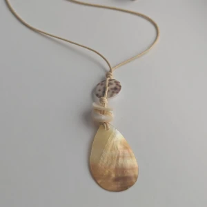 Halsband med hängsmycke i guld och pärlemor - Ett elegant halsband med ett hängsmycke i guld och pärlemor. Halsbandet har en tunn rem och hängsmycket består av en droppformad guldfärgad platta, en rund pärlemorplatta med bruna fläckar och flera små vita pärlor.