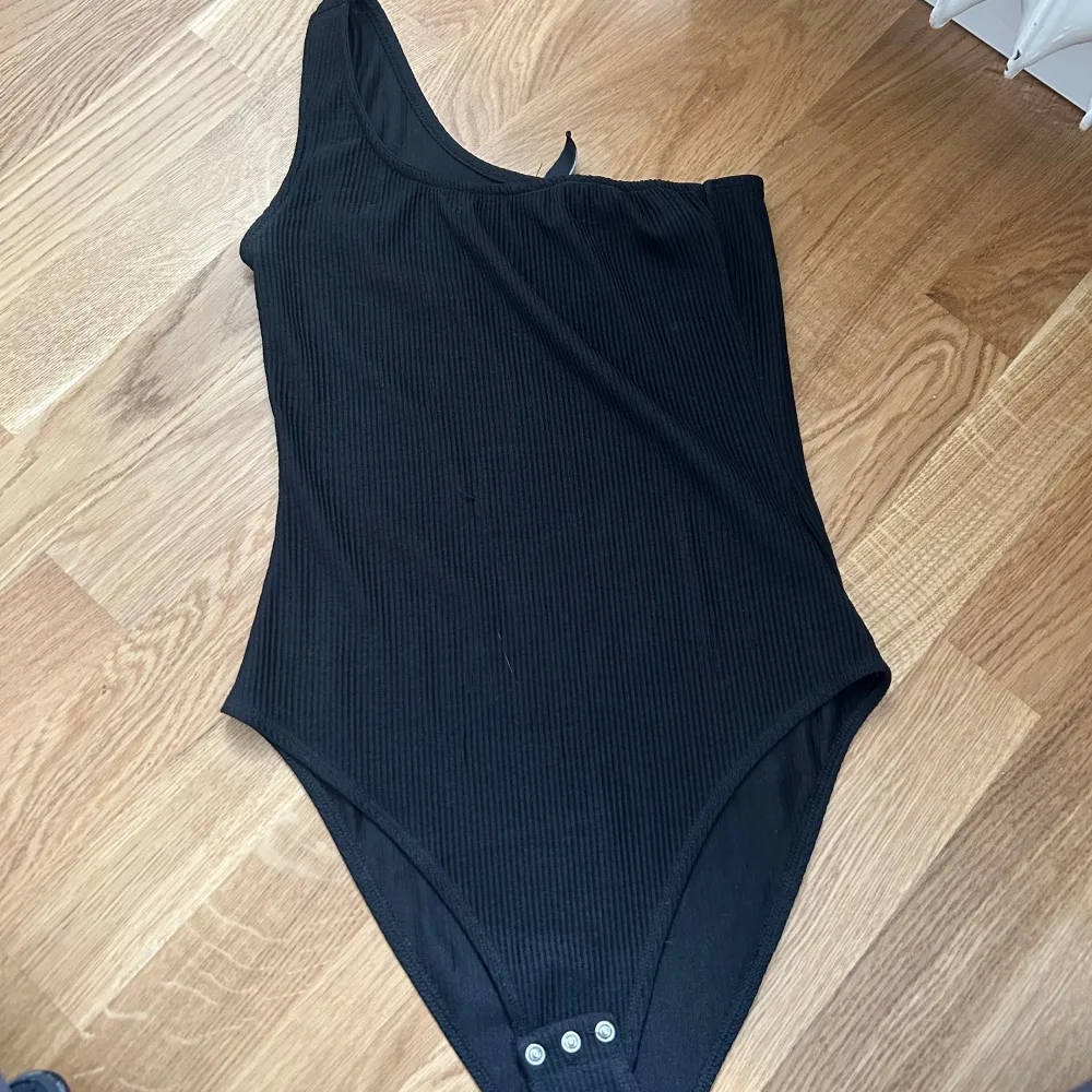 En svart ribbad oneshoulder bodysuit med knappar i grenen. Bodysuiten har en asymmetrisk design med en axelbandslös sida.. Toppar.