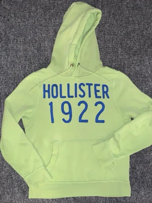 Ljusgrön hoodie från Hollister - Snygg ljusgrön hoodie från Hollister med stor blå text 'HOLLISTER 1922' på bröstet. Klassisk huva med dragsko och stor magficka framtill. Perfekt för en avslappnad och trendig look. Tillverkad i mjukt material för extra komfort.