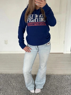 Blå UFC hoodie med tryck - Säljer en blå hoodie med texten 'Ultimate Fighting Championship' framtill och 'ROOT LEE 99' på ryggen. Hoodien har huva och magficka, samt vita och röda tryck. Perfekt för dig som gillar sportig stil och vill sticka ut.