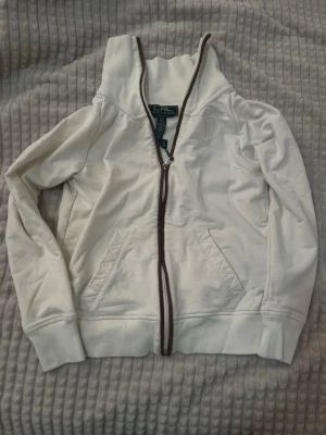 Vit kofta från Lauren Ralph Lauren - Snygg vit hoodie från Lauren Ralph Lauren med brunt kantband längs dragkedjan. Broderad logga på bröstet och två fickor framtill. Tillverkad i bomull och polyester med lite stretch för extra komfort. Perfekt för en chill och stilren look.