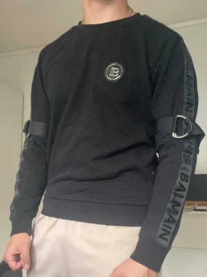 Svart sweatshirt med detaljer från Balmain Paris - Cool svart sweatshirt från Balmain med stora D-ringar och band på ärmarna. Logga på bröstet och texten 'BALMAIN' längs båda ärmarna. Tröjan har rund hals och ribbade muddar. Perfekt för dig som gillar streetwear och unika detaljer.