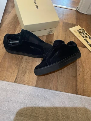 Svarta sneakers från Axel Arigato - Stilrena svarta sneakers från Axel Arigato i mocka med diskret logga på sidan. Skorna har en chunky svart sula och minimalistisk design utan synliga snören. Perfekta för dig som gillar en clean och modern look.