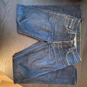 Mörkblå jeans från Nudie Jeans - Säljer ett par klassiska mörkblå jeans från Nudie Jeans med raka ben och kontrastsömmar. Jeansen har fem fickor, gylf med dragkedja och Nudie-logga på bakfickan samt läderpatch i midjan. Perfekta till din vardagsstil!