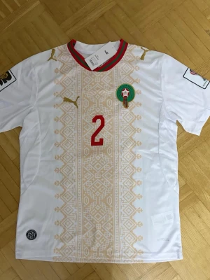 Marocko fotbollströja Hakimi Puma - Säljer en vit Marocko fotbollströja från Puma med Hakimi och nummer 2 på ryggen. Tröjan har gulddetaljer, rött tryck och ett traditionellt mönster framtill. Marockos landslagsemblem på bröstet och korta ärmar. Perfekt för match eller träning.