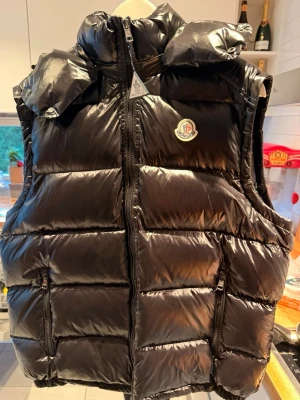  Moncler väst  - Snygg svart dunväst från Moncler med glansig finish och avtagbar huva. Västen har två dragkedjeförsedda fickor framtill och Moncler-logga på bröstet. Perfekt för lager-på-lager och håller dig varm under kyliga dagar.