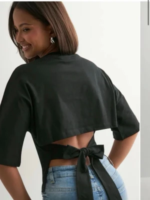 Svart topp med öppen rygg och knyt - Trendig svart topp med öppen rygg och breda knytband i midjan. Toppen har halvlånga ärmar och en croppad passform som ger en cool och modern look. Perfekt att styla med höga jeans eller kjol för en snygg outfit.
