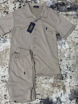 Beige set från Polo Ralph Lauren - Snyggt beige set från Polo Ralph Lauren med kortärmad skjorta och matchande shorts. Skjortan har knappar framtill, klassisk krage och mörkblå logga på bröstet. Perfekt för en avslappnad look.