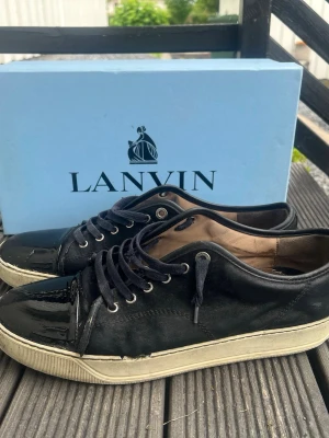 Svarta Lanvin sneakers med lackdetaljer - Snygga svarta sneakers från Lanvin med glansiga lackdetaljer på tån och hälen. Skorna har svarta skosnören, vita sulor och en klassisk låg profil. Materialmix av mocka och lack ger en lyxig känsla och unik look.