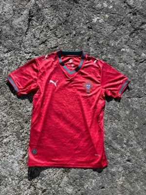 Röd Portugal fotbollströja Puma - Snygg röd Portugal fotbollströja från Puma med gröna detaljer vid krage och ärmslut. Tröjan har korta ärmar, V-ringning och Portugal-emblem på bröstet. Tillverkad i lätt och ventilerande material, perfekt för träning eller match. Vid frågor hör av er