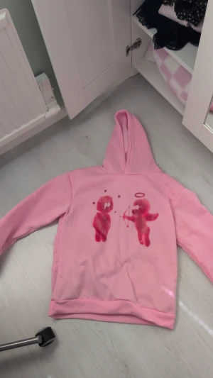 Rosa hoodie med grafiskt tryck - Säljer en ljusrosa hoodie med huva och ett grafiskt tryck framtill som föreställer två figurer, varav en med änglavingar och pilbåge. Hoodien har en avslappnad passform och ribbade muddar vid ärmar och nederkant. Perfekt för dig som gillar unika och lekfulla plagg.