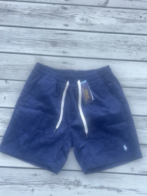 Marinblå manchester shorts Polo Ralph Lauren - Säljer ett par marinblå manchester shorts från Polo Ralph Lauren med vit broderad logga på benet och vita snören i midjan. Shortsen har avslappnad passform och sidofickor. Perfekta för sommardagar och chill vibes.