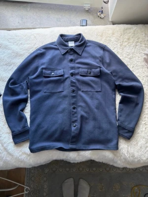 Blå overshirt från Selected Homme - Snygg blå overshirt från Selected Homme med klassisk krage och två bröstfickor med knappar. Overshirten har långa ärmar och stängs med knappar framtill. Perfekt att bära över en t-shirt för en avslappnad men stilren look.