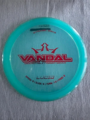 Turkos disc Vandal Lucid - En turkos disc från Dynamic Discs, modell Vandal Lucid. Discen har röd text och grafik, är rund och tillverkad i plast. Perfekt för dig som gillar discgolf och vill ha en understabil fairway driver.