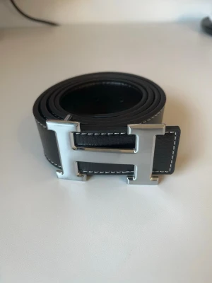 Svart läderbälte med H-spänne - Snyggt svart bälte i läder med ett stort, silverfärgat H-format spänne. Bältet har vita kontrastsömmar längs kanterna som ger en stilren look. Perfekt accessoar för att lyfta din outfit.