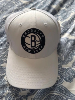 Vit Brooklyn NBA keps - Snygg vit keps med Brooklyn Nets-logga framtill och NBA-märke baktill. Klassisk böjd skärm och justerbar passform. Tillverkad i ett ventilerande material som passar perfekt till streetwear-stilen. Liten sminkfläck som går bort i tvätten
