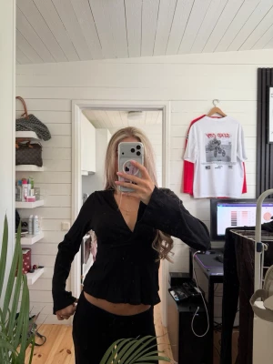 Svart omlottblus med v-ringning - Snygg svart omlottblus med djup v-ringning och långa ärmar. Blusen har knytband i midjan som ger en feminin siluett och lätt utsvängd nederkant. Perfekt att matcha med både jeans och kjol för en trendig look.