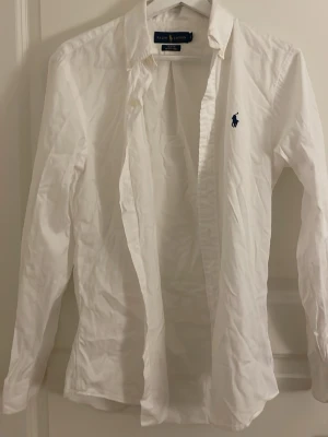 Vit skjorta från Ralph Lauren - Klassisk vit skjorta från Ralph Lauren med blå logga broderad på bröstet. Skjortan har lång ärm, knappar framtill och är tillverkad i bomull. Perfekt till många olika stilar och enkel att matcha.