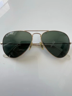 Ray-Ban aviator solglasögon guld 55mm - Klassiska Ray-Ban aviator solglasögon 55 mm med guldfärgad metallbåge och mörkgröna glas. Snygg och tidlös modell som passar till det mesta. Tunn ram och bekväma näskuddar för extra komfort. Passar perfekt för dig med litet eller lite smalare ansikte👌