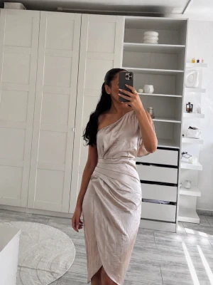 Champagnefärgad omlottklänning oneshoulder - Snygg champagnefärgad klänning med omlottkjol och oneshoulder-design. Klänningen har draperade detaljer och en kort ärm på ena sidan, vilket ger en unik och trendig look. Perfekt för dig som vill sticka ut med stilrena detaljer.