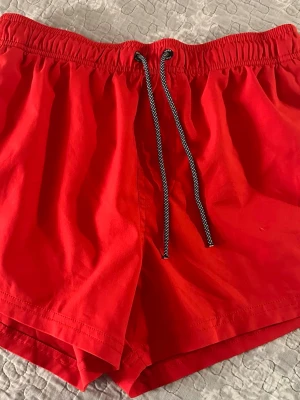 Röda shorts med snörning S - Snygga röda shorts med elastisk midja och svartvit snörning framtill. Shortsen har en bakficka och är tillverkade i ett lätt och bekvämt material, perfekta för varma dagar.