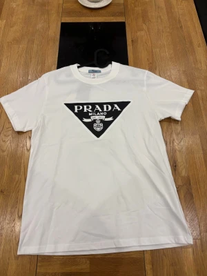 Prada tshirt - Snygg vit t-shirt med stort svart triangulärt Prada Milano-tryck på bröstet. Klassisk rund hals och korta ärmar. Perfekt för dig som gillar stilrena och trendiga plagg med tydlig logga. Tillverkad i mjuk bomull för skön känsla.