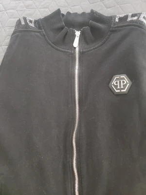 Philipp plein Svart tracksuit med dragkedja som en L - Snygg svart tracksuit med dragkedja framtill och matchande byxor. Setet har vita detaljer, ribbade muddar och PP-logga på både jacka och byxa. Axelpartiet och sidan på byxorna har ett mönstrat band med text. Perfekt för chill eller streetwear. Som en större lagre i storlek 