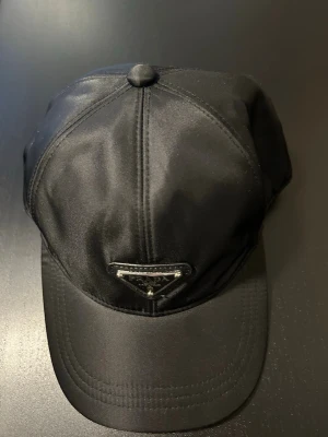 Svart keps från Prada i nylon - Snygg svart keps från Prada med klassisk böjd skärm och det ikoniska metalltriangel-logotypen framtill. Tillverkad i glansigt nylonmaterial som ger en modern och exklusiv känsla. Perfekt accessoar för en trendig streetwear-look.