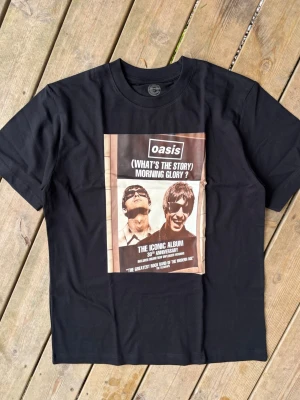 Svart Oasis band t-shirt - Svart t-shirt med tryck av Oasis och albumet '(What's The Story) Morning Glory?' på framsidan. Motiv med två personer och text om albumets 30-årsjubileum. Rund hals och korta ärmar. Perfekt för dig som gillar klassisk britpop.