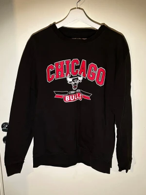 Svart Chicago Bulls sweatshirt - Cool svart sweatshirt med Chicago Bulls-tryck i rött och vitt framtill i storlek M. Tröjan har rund halsringning och långa ärmar. Perfekt för dig som gillar sportig streetstyle och vill visa ditt basketintresse. Är i allmänt bra skick men har en  slitning på trycket där storlek brukar stå.