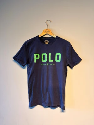 Blå POLO t-shirt från Ralph Lauren - Mörkblå t-shirt från Ralph Lauren med stort grönt POLO-tryck och logga på bröstet. Klassisk rund hals och korta ärmar. Tillverkad i mjuk bomull för en skön och avslappnad stil.