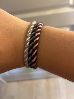 Randigt vänskapsarmband i pastell - Ett handgjort vänskapsarmband i tvinnad design med ränder i ljusblått, svart, vitt och rosa. Armbandet är gjort av mjukt garn och har en enkel, avslappnad stil som passar perfekt till vardagslooken. Säljs styckvis och är lätt att matcha med andra accessoarer.