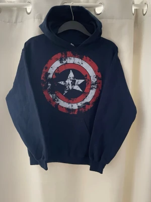 Mörkblå hoodie usa tryck  - Tja! Snygg mörkblå hoodie med stor usa sköld/captain america sköld på bröstet i rött, vitt och blått. Hoodien har huva och magficka, samt ribbade muddar vid ärmar och nederkant. Perfekt för dig som gillar denna stil och vill ha en bekväm och stilren tröja. Passar perfekt till allt och allmänt riktigt snygg!! Skick 9/10 och storlek S! Hör av dig vid mer frågor eller funderingar🕺mvh Ga😁🤩