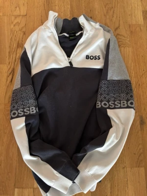 Grå och vit half zip tröja från BOSS - Säljer en snygg grå och vit tröja från BOSS med half zip och tryckt logga på bröstet samt armarna. Tröjan har lång ärm och en sportig look med kontrastpaneler. Perfekt för dig som gillar streetwear och vill ha något stilrent. Pris kan diskuteras vid snabb affär!