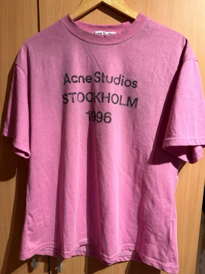 Rosa Acne Studios t-shirt - Säljer en rosa t-shirt från Acne Studios med tryck 'STOCKHOLM 1996' på bröstet. T-shirten har rund hals, korta ärmar och en relaxed passform. Perfekt för dig som gillar streetwear och vill ha något stilrent men ändå statement.