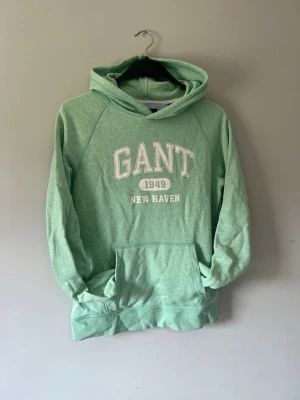 Mintgrön hoodie från GANT - Snygg mintgrön hoodie från GANT med tryck 'GANT 1949 NEW HAVEN' på bröstet. Klassisk känguruficka framtill och huva. Tillverkad i mjukt bomullsmaterial, perfekt för en avslappnad och sportig stil.