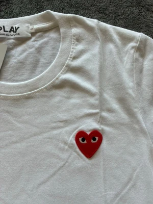 Comme des Garçons T-shirt  - Vit t-shirt från Comme des Garçons PLAY med det ikoniska röda hjärtat med ögon broderat på bröstet. Klassisk rund halsringning och korta ärmar. Tillverkad i mjuk bomull för en skön och clean look. Storlek S. Vid snabb affär kan priset diskuteras 