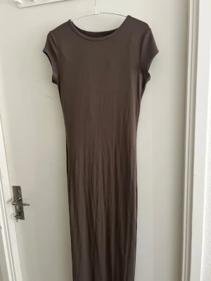 Brun långklänning med kort ärm - En enkel och stilren brun långklänning med rund halsringning och korta ärmar. Klänningen har en rak passform och är tillverkad i ett mjukt, stretchigt material som ger en bekväm känsla. Perfekt för dig som gillar minimalistisk stil.