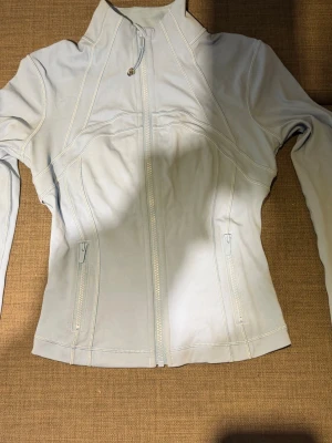 Ljusblå define jacket från Lululemon - Snygg ljusblå zip-jacka från Lululemon med hög krage och figurnära passform. Jackan har två fickor med dragkedja och dekorativa sömmar som ger en sportig look. Perfekt till träning eller som ett extra lager. Croppad model, sista bilden ser man färgen bättre. Storlek 4. Har dragit bort lappar men har kvitto