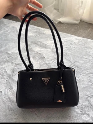Svart handväska från Guess - Stilren svart handväska från Guess med dubbla långa handtag och guldfärgade detaljer. Väskan har en klassisk rektangulär form, Guess-logga framtill och en dekorativ tag. Tillverkad i slätt konstläder med dragkedja upptill.