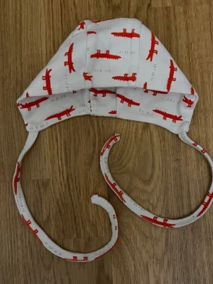 Vit barn / baby mössa med röda krokodilmotiv, storlek 3-6 månader - Söt och praktisk vit babymössa med röda krokodilmotiv och knytband som hjälper mössan att sitta på plats! Storlek: 3–6 månader. Säljs i bra skick! Kika gärna in på mina andra annonser, jag säljer just nu flera barn- och babyplagg samt barnartiklar! Hör av dig vid intresse och frågor!