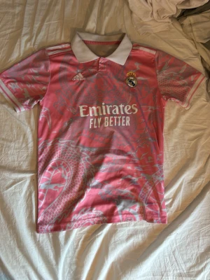 Rosa Real Madrid tröja Adidas - Säljer en rosa Real Madrid matchtröja från Adidas med vitt krage och knapp. Tröjan har ett drakmönster i ljusrosa och vitt, samt Real Madrid-logga och Adidas-logga på bröstet. Trycket 'Emirates Fly Better' på framsidan. Perfekt för fotbollsfans!